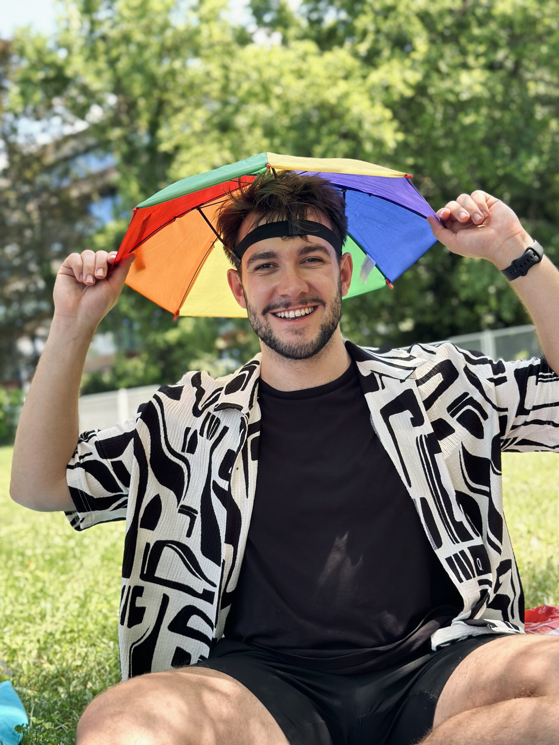 Adrian smiling sitting on grass wearing a rainbow hat